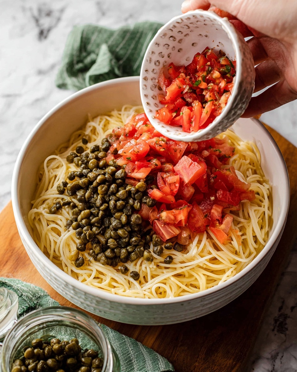 Lemon Capellini Salad Recipe 4 A white bowl filled with a base layer of light yellow cooked spaghetti. On top of the spaghetti, there are two distinct topping sections: one side has dark green capers with a round, slightly shiny texture, and the other side has bright red diced tomatoes with a fresh, soft appearance. A woman's hand is holding a small white bowl with dimples and is pouring more diced tomatoes into the bowl. The bowl is placed on a white marbled surface with a green cloth nearby and a jar of capers in the foreground. photo taken with an iphone --ar 4:5 --v 7