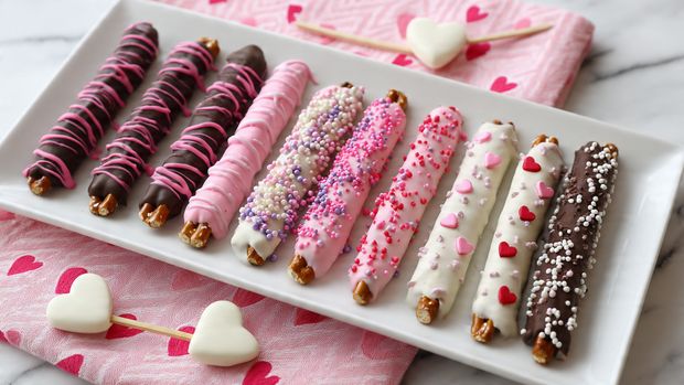 A long white rectangular plate holds eight chocolate-covered pretzel rods arranged in a neat row on a white marbled surface with a pink heart-patterned cloth beneath part of the plate. The first two pretzels are coated in dark chocolate with bright pink drizzles, the next two are white chocolate covered with small pink and purple heart sprinkles. The following two rods are bright pink coated with white and pink round sprinkles. The next two rods are white chocolate covered with pink drizzles and tiny heart decorations. The last two rods are covered in milk chocolate with white, pink, and red sprinkles. The pretzel ends are left uncovered on all rods. Three white heart-shaped objects on sticks rest beside the plate. photo taken with an iphone --ar 4:5 --v 7