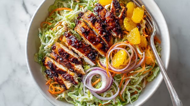 A white bowl is filled with a colorful salad base made of thinly sliced light green cabbage, orange carrot strips, light purple onion slices, and small pieces of bright yellow orange segments scattered throughout. On top, there are several pieces of grilled chicken with a shiny, dark brown, and slightly charred glaze. A few black sesame seeds and small green herbs are sprinkled over the salad. A golden fork rests on the right side of the bowl. The bowl sits on a white marbled surface. photo taken with an iphone --ar 4:5 --v 7