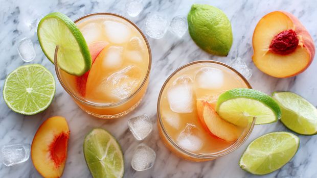 Two round glasses filled with light orange juice and ice cubes sit on a white marbled surface. Each glass is topped with a wedge of lime on the right side and a peach slice on the left side, both resting on the edge. Around the glasses are scattered ice cubes, lime halves and wedges, and peach slices. The colors include the light orange liquid, bright green lime, and warm orange peach against the white marbled background. The photo taken with an iphone --ar 4:5 --v 7