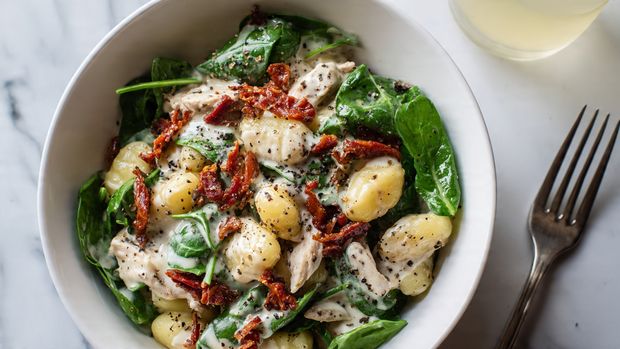 A pan filled with creamy gnocchi mixed with wilted green spinach leaves and halved bright red cherry tomatoes, topped with golden crispy breadcrumbs and a light sprinkling of shredded white cheese. The gnocchi are small, soft, and plump with a smooth yellowish texture, sitting in a thick light yellow cream sauce. A wooden spoon rests inside the pan, partially lifting some gnocchi, showing the richness and texture of the sauce and toppings. The pan is on a white marbled surface with a white cloth with red stitching nearby. Photo taken with an iphone --ar 4:5 --v 7