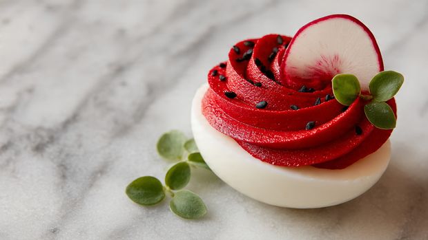 The image shows a round white plate filled with two layers of deviled eggs arranged neatly. Each egg half has a smooth white outer layer with a thick pink creamy filling layered inside. On top of the pink filling, there is a small green herb garnish, and a tiny sprinkle of seasoning adds a slight texture and darker color contrast. The plate is set on a white marbled surface. Photo taken with an iphone --ar 4:5 --v 7