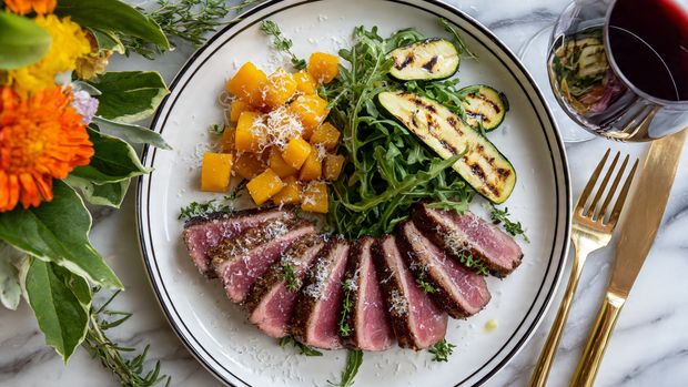 The dish features five slices of medium-rare pink meat arranged in a fan shape on the right side of a white plate with a thin black ring around its edge, garnished with small sprigs of herbs and thin garlic slices. In the center sits a small pile of fresh dark green arugula leaves, topped with several grilled yellow-green zucchini slices. On the left side, there is a neat mound of bright orange roasted cubes, possibly sweet potatoes, sprinkled with fine grated cheese. The plate is placed on a white marbled surface, with a gold fork and knife on the right and a glass of red wine in the top right corner. A woman's hand holding a fork is partially visible near the top left corner, beside green leaves and vibrant orange flowers. Photo taken with an iphone --ar 4:5 --v 7
