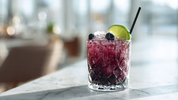 A clear glass filled with a light green drink containing multiple bright green mint leaves layered throughout the liquid, with some mint leaves near the bottom and others floating near the top mixed with ice cubes. A thin, translucent slice of lime is placed on the rim of the glass, and a sprig of fresh vibrant mint sticks out slightly above the rim. The glass sits on a white marbled surface with a blurred natural light background. photo taken with an iphone --ar 4:5 --v 7
