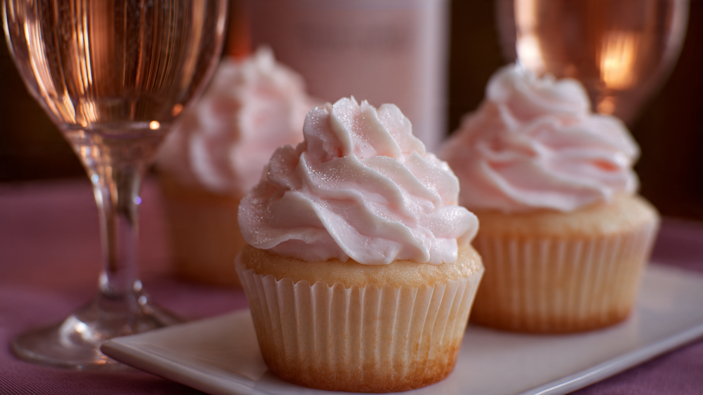 A group of eight light golden cupcakes sit on a white plate, arranged in close rows. Each cupcake is topped with a thick, smooth layer of pale pink frosting that is piped in two different styles: some have soft, rosette swirls with delicate ridges, and others have simple, flat spirals with a creamy texture. The cupcake in the front center has its white wrapper opened and peeled down, showing a super soft and moist crumb inside. The whole scene is set against a background of green leaves, with lighting that highlights the smooth frosting and soft cake textures. photo taken with an iphone --ar 4:5 --v 7