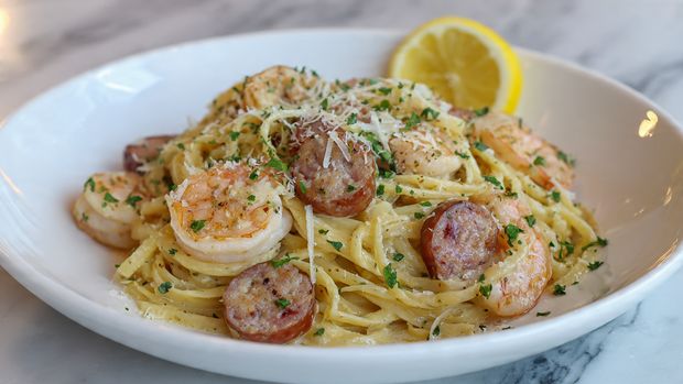 The image shows a white bowl filled with spaghetti pasta mixed with several grilled shrimp. The shrimp are golden-brown with slight char marks, giving a textured look. The creamy sauce coats the spaghetti, showing a light orange color speckled with red bits, possibly from spices or small pieces of tomato. There are small green herb pieces scattered across the dish, adding contrast. White grated cheese is sprinkled unevenly on top, adding texture and brightness. A silver fork is twirling some spaghetti on the right side of the bowl. The bowl sits on a white marbled surface. Photo taken with an iphone --ar 4:5 --v 7
