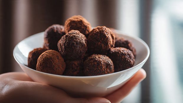 A white plate filled with round chocolate-covered balls stacked closely together. Each ball has a smooth, shiny milk chocolate coating with a slightly uneven texture, revealing bits of a crunchy golden brown inside at the base. The balls are arranged in several layers, creating a small mound. The white marbled surface below the plate adds a clean, simple background. photo taken with an iphone --ar 4:5 --v 7