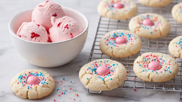 A white bowl with five light pink ice cream scoops, each having a soft texture and small red bits inside, sits on a white marbled surface. Around the bowl are several round white cookies on a metal cooling rack; some cookies have a smooth light beige color, others are coated with small pastel sprinkles in blue, pink, yellow, and white. Each cookie has a shiny light pink candy piece shaped like a small peak in the center. In the background, a white waffle cone and a few scattered pink candies are visible on the white marbled surface. photo taken with an iphone --ar 4:5 --v 7