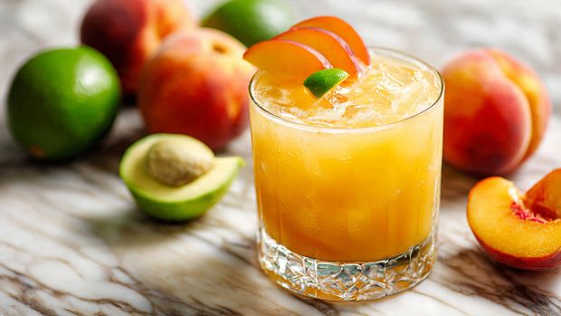 A round clear glass filled with a light orange drink with ice cubes inside, topped with two thin slices of peach and a small green lime wedge placed on the ice near the rim. The drink looks smooth and slightly frothy on the surface. The glass is set on a white marbled surface with fresh whole and sliced peaches, a green lime, and a green avocado around it, softly blurred in the background. The lighting is bright and natural, highlighting the fresh colors and textures. photo taken with an iphone --ar 4:5 --v 7
