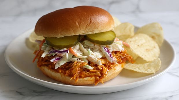 A white plate on a white marbled surface holds a sandwich with a soft, shiny golden bun as the top layer. Below the bun are three green pickle slices, followed by a layer of creamy coleslaw with visible purple and white cabbage strands. Under the coleslaw is a thick layer of shredded chicken mixed with glossy orange barbecue sauce. Next to the sandwich, a small pile of light golden potato chips is placed on the plate. In the background, a slightly blurred second sandwich and a small clear bowl with pickle slices are visible. photo taken with an iphone --ar 4:5 --v 7
